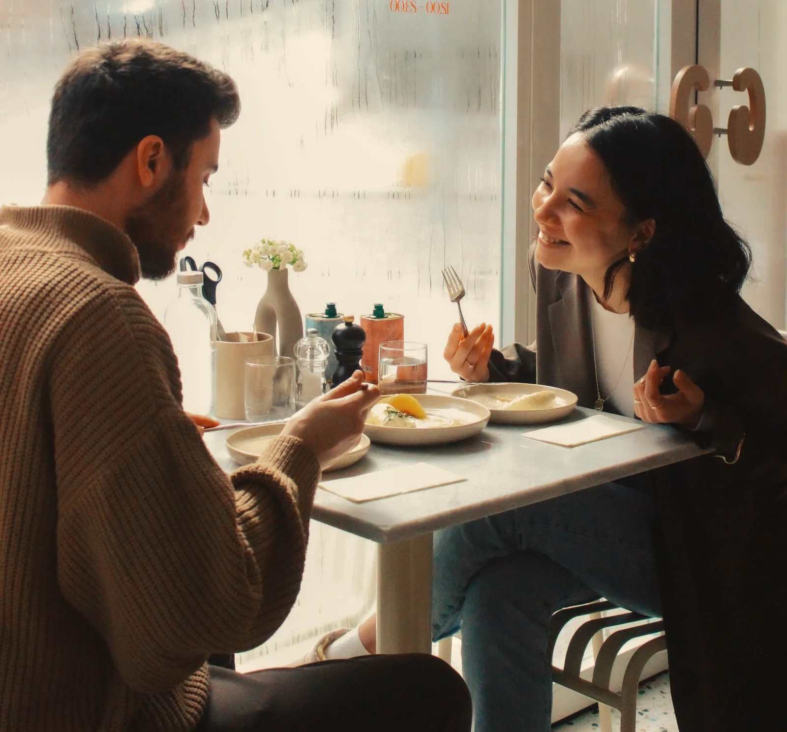 Two people at a small café table by a big window, one of them smiling at the other over lunch
